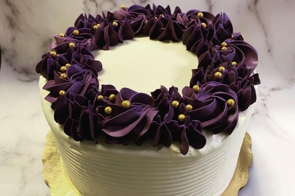 Round white buttercream cake with a ring of purple piped rosettes and gold pearls on a gold cake board