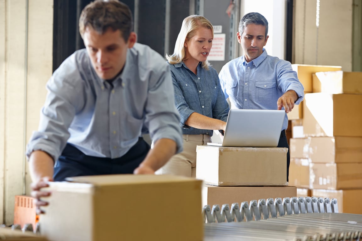 Three warehouse workers collaborating with one using a laptop on top of a cardboard box along a conveyor belt.
