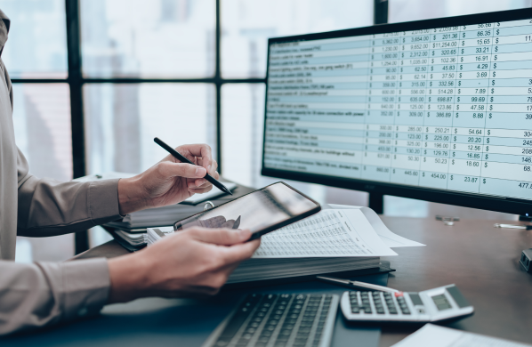 An individual analyzing complex financial data across multiple computer screens, with a focus on a spreadsheet.