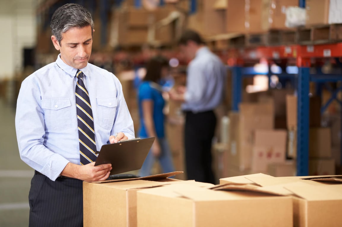 Warehouse manager in a blue shirt and tie reviewing a clipboard over a cardboard box with shelves and workers in the background.