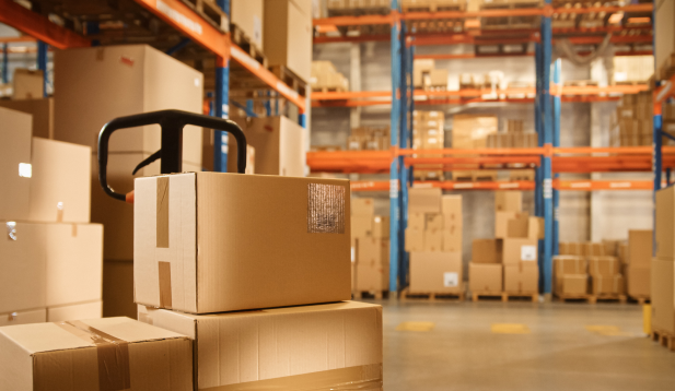 Close-up view of cardboard boxes stacked on a hand dolly in a warehouse with shelves and boxes in the background.
