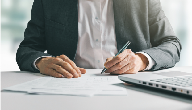 Close-up of a person's hands holding a pen over a document next to a laptop on a desk.