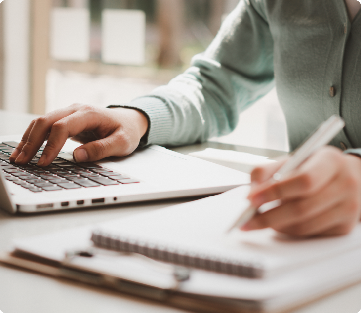A person's hands typing on a laptop keyboard while taking notes with a pen on a notepad.
