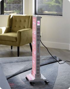 Vital Red LIght. Red Light Therapy. A vertical red light therapy panel on wheels is plugged in and illuminated in a modern living room near a green armchair and a window.