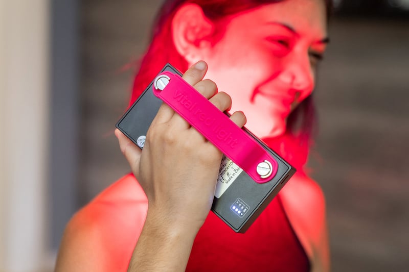 Vital Red LIght. Red Light Therapy. Person holding a rectangular device labeled "vital red light," emitting red light onto their face, while a Wholesale Dashboard appears blurred in the background.