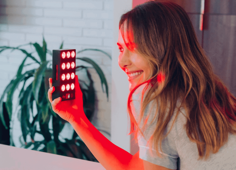 A woman smiles while holding a handheld red light therapy device with multiple LEDs. She is indoors near a plant and a white brick wall, harnessing the vital charge of this innovative gadget to enhance her well-being.