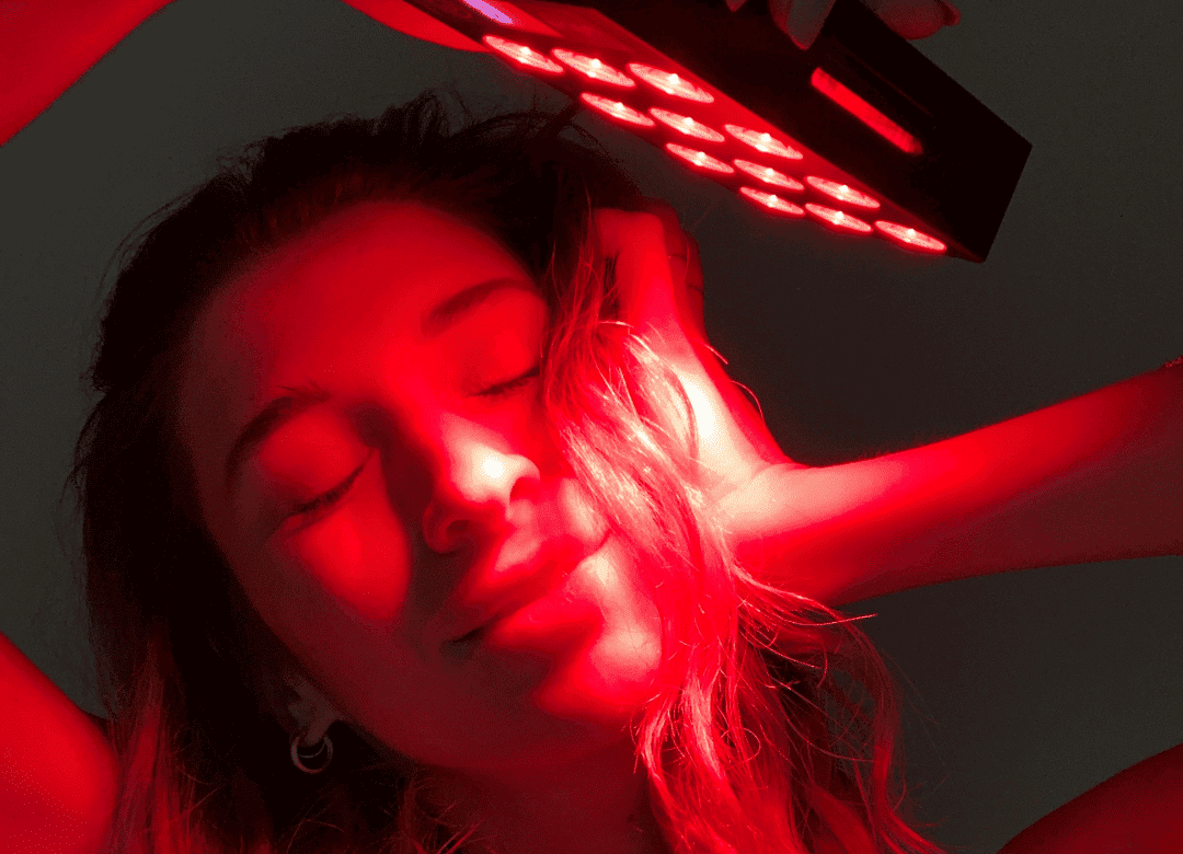 A person with closed eyes holds a handheld red light therapy device above their head, basking in the vital charge of its illumination.