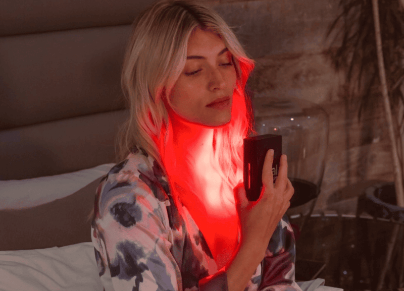 Vital Red LIght. Red Light Therapy. A person sits on a bed, eyes closed and wearing a patterned robe, holding a handheld red light therapy device emitting a vital charge against their neck.