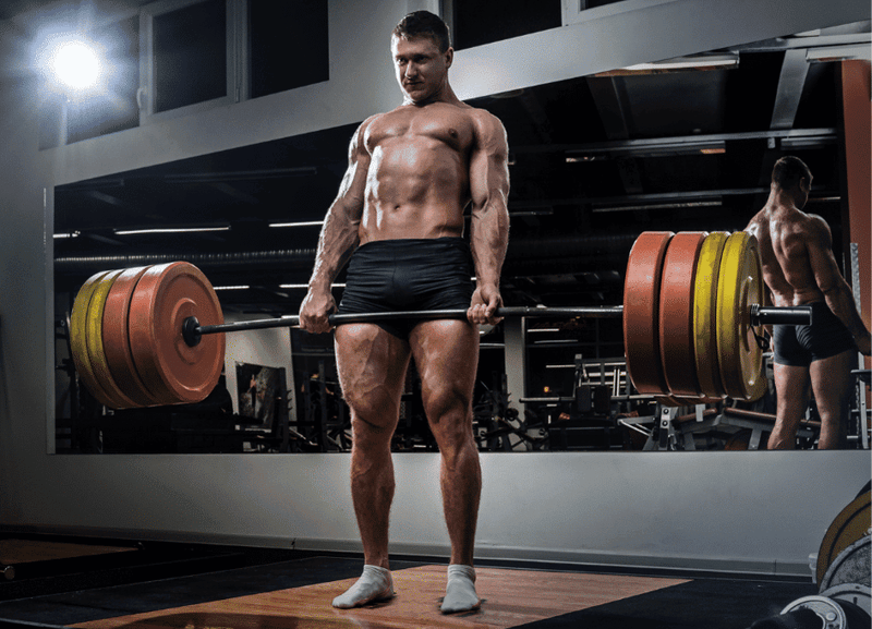 A muscular person performs a deadlift with a heavily loaded barbell in a gym, standing on a wooden platform. A large mirror and gym equipment are visible in the background, highlighting the dedication put into their fitness journey—perhaps boosted by the benefits of red light therapy for recovery.