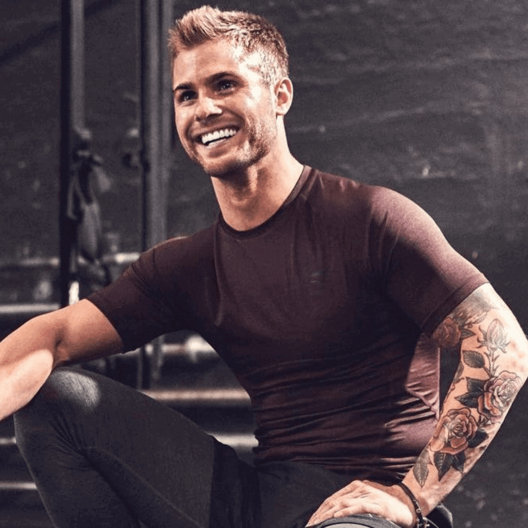 A person with short hair and a tattoo on their left arm smiles while crouching in a gym setting. They're wearing a fitted, short-sleeve T-shirt and pants, clearly benefiting from the red light therapy that keeps their energy high for workouts.