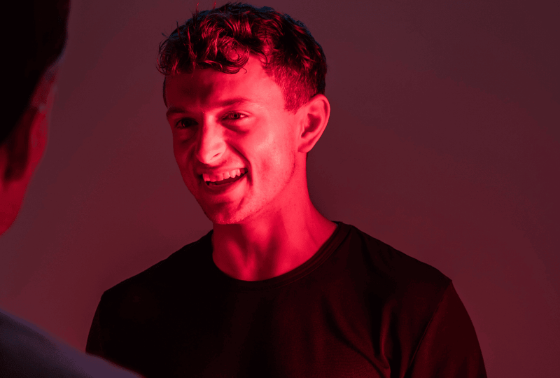 A person smiling under red lighting, donning a dark shirt, faces another who is partially visible, capturing the essence of connection. This moment could be part of an "about us" section, illustrating the warmth and camaraderie within our team.