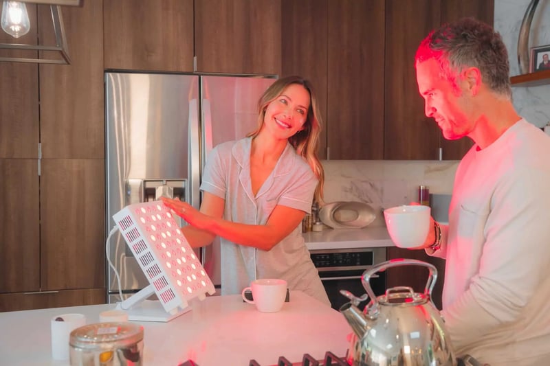 A woman and man stand in a kitchen, their smiles radiant like the red light therapy device glowing on the counter beside them. They each hold steaming mugs, basking in both warmth and wellness.