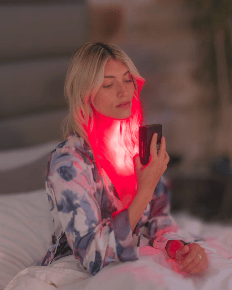 Person with closed eyes holds a glowing red light panel to their neck, sitting on a bed and wearing a patterned robe.