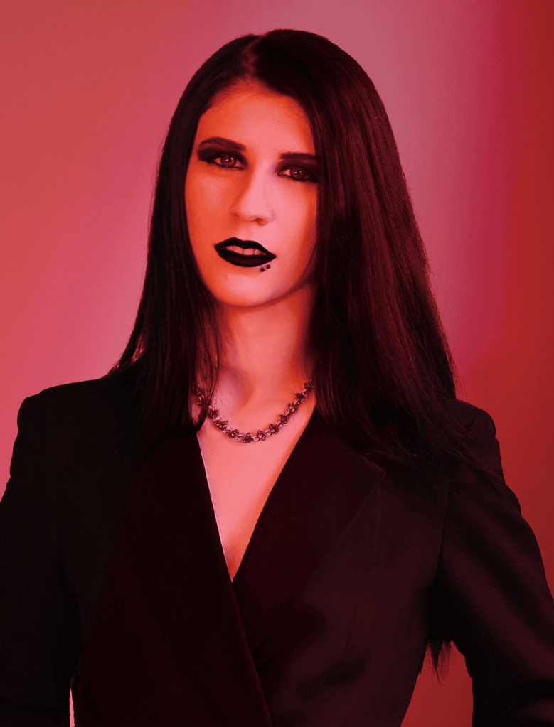 About us: A person with long dark hair and dramatic makeup wears a black blazer and necklace, set against a vivid red-toned background.