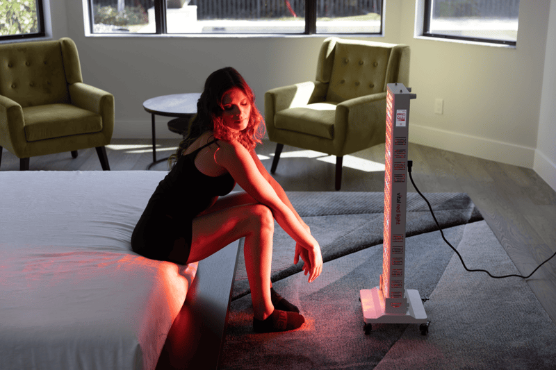 A woman sits on the edge of a bed in a sunlit room, facing a standing Vital Pro 2.0 red light therapy device that is turned on.