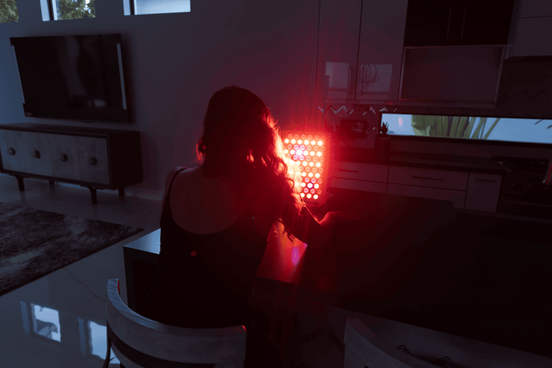 A person sits at a kitchen counter in a dimly lit room, facing the bright glow of a Vital Pro 2.0 red light therapy panel.