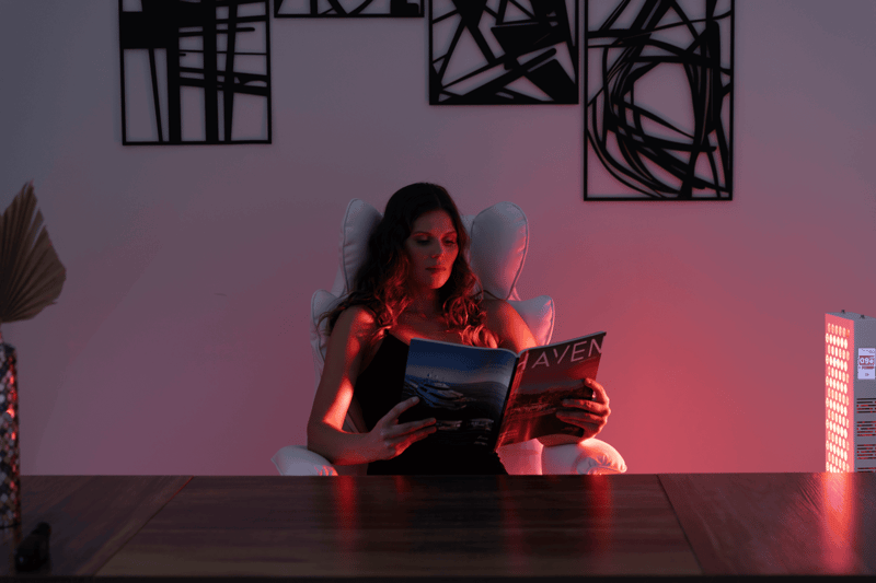 A woman sits in a white chair reading a magazine at a wooden desk, illuminated by pink and purple lighting from the Vital Pro 2.0, with abstract art on the wall behind her.