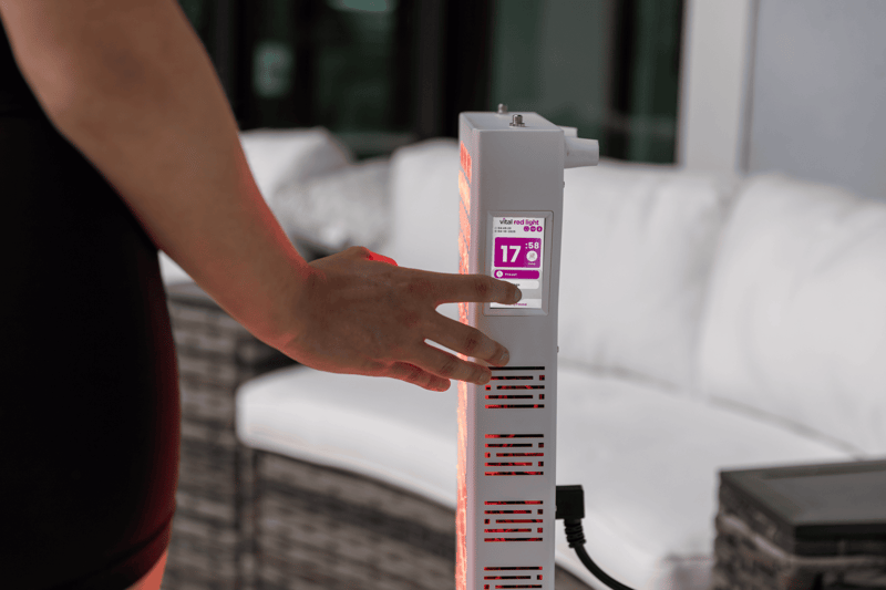 A person presses a button on the Vital Pro 2.0 electronic device with a digital display reading "17:00" near a white outdoor sofa set.