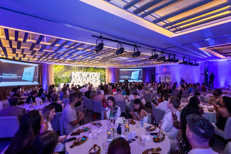 Guests sit at elegantly adorned tables, eagerly awaiting the night's festivities. The venue gleams with vibrant decor, while a prominent sign in the background proudly displays "NEW YOU AWARDS," setting the stage for a memorable celebration of achievements.