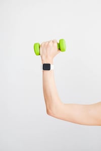 A hand wearing a smartwatch holds a small green dumbbell against a plain white background biohacking health
