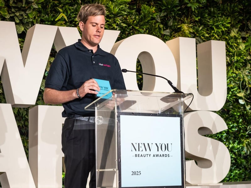 A speaker stands at the podium, framed by a vibrant green leafy background and large white letters announcing the prestigious "New You Beauty Awards 2023.