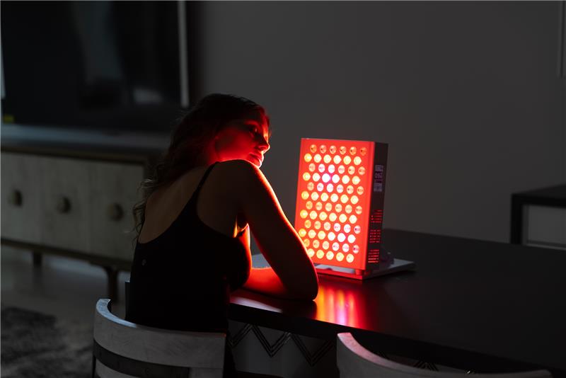 Vital Red LIght. Red Light Therapy. A person sits at a table facing a red light therapy device, which is turned on and emits bright red light onto their face and upper body. Book a consultation to experience the benefits of red light therapy for yourself.