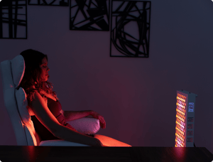 Vital Red LIght. Red Light Therapy. A woman sits in a chair facing a panel of red and yellow lights in a dimly lit room with abstract wall art, reminiscent of modern chiropractic clinics.