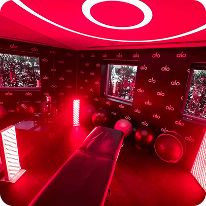 Vital Red LIght. Red Light Therapy. Room with red lighting, a massage table, exercise balls, LED red light panels, and "alo" logos on the walls; two windows show outdoor greenery.