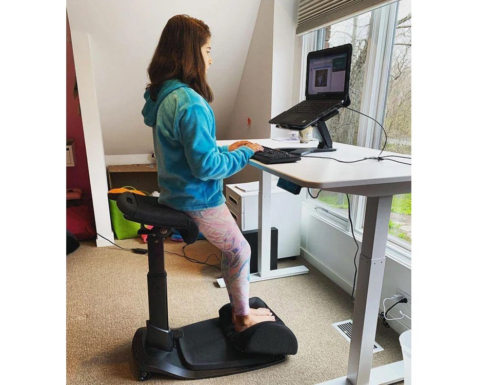 TechDogs-"Ergonomic Chairs"-"Image Of Young Woman Using A Kneeling Chair And Standing Desk Setup While Working On A Laptop In A Bright Home Office"