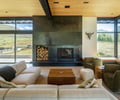 Preview of Sun Valley Fireweed - Image 24: floor-to-ceiling windows, wooden ceiling, modern fireplace, built-in wood storage, beige sofa, glass walls, natural light, open view, wooden coffee table, rustic decor, greenery outside, comfortable seating, minimalist design, lounge area, contemporary style.