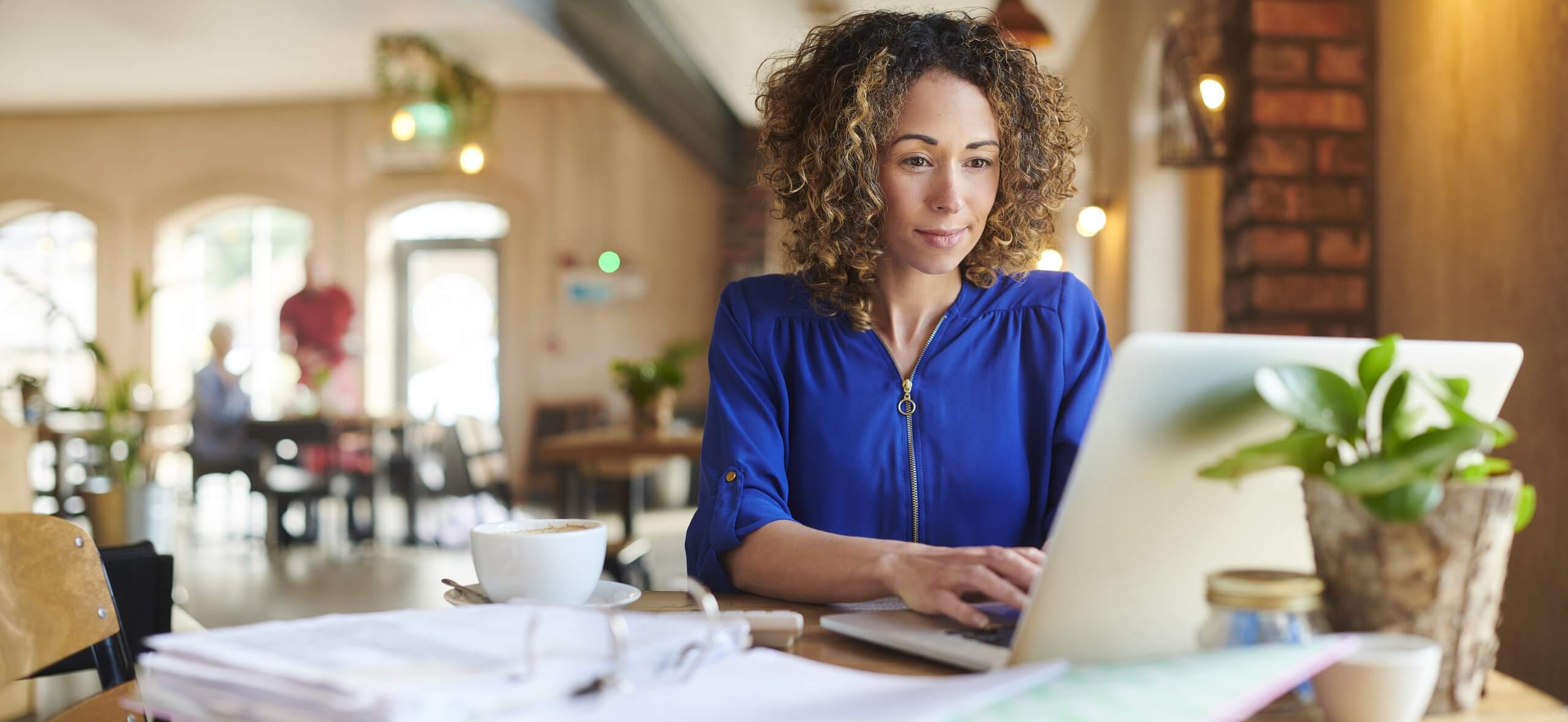 Woman with curly hair and blue shirt works on a laptop at a cafe - prioritizing.