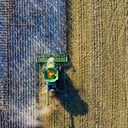 An aerial view of a green combine harvester cutting a clean diagonal line across a field, harvested stubble on one side and standing crop on the other.