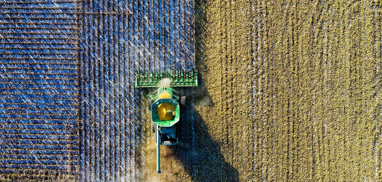 An aerial view of a green combine harvester cutting a clean diagonal line across a field, harvested stubble on one side and standing crop on the other.