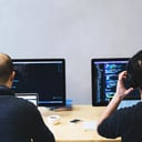 Two developers seen from behind at a shared desk, each working at a monitor filled with code; one is wearing headphones.