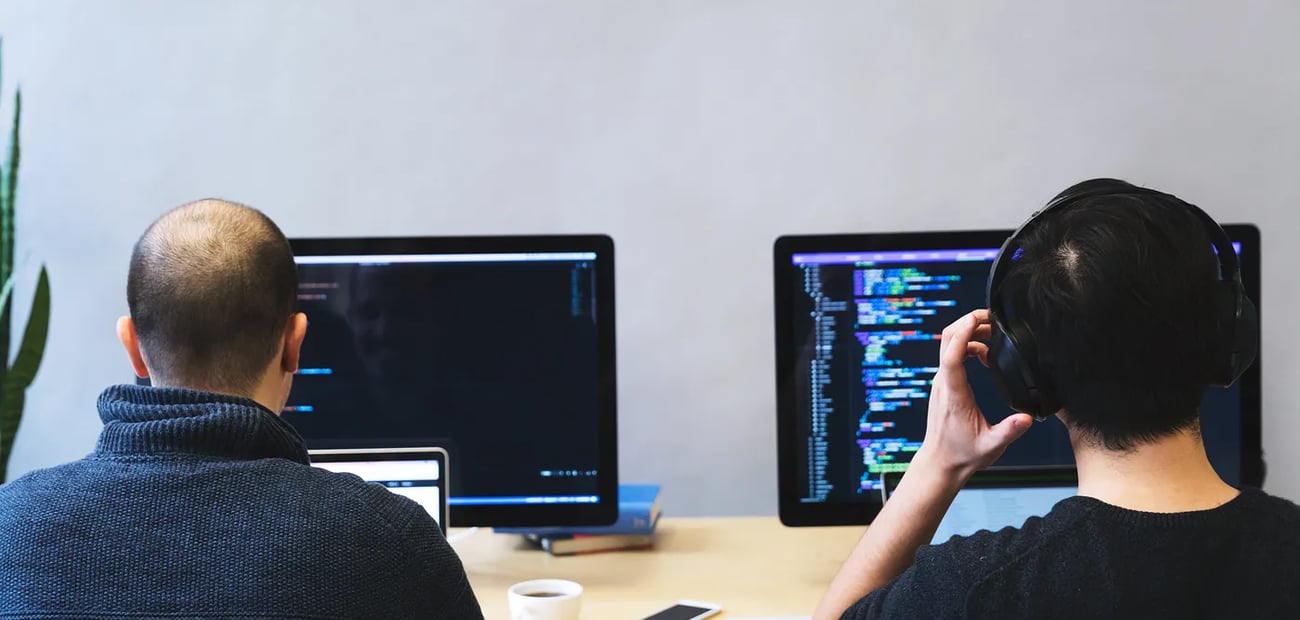 Two developers seen from behind at a shared desk, each working at a monitor filled with code; one is wearing headphones.
