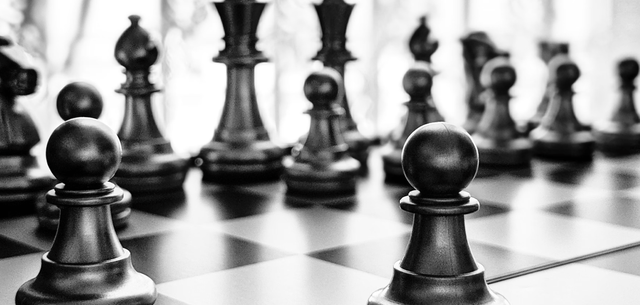 A black-and-white photograph of a chess board mid-game, a single pawn sharp in the foreground and the king and queen out of focus behind it.