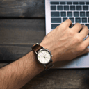 A wrist wearing a brown leather chronograph watch, resting over a laptop keyboard on a dark wooden desk.