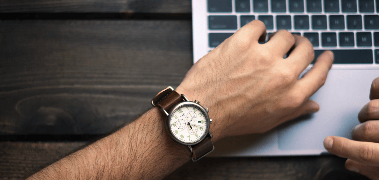 A wrist wearing a brown leather chronograph watch, resting over a laptop keyboard on a dark wooden desk.