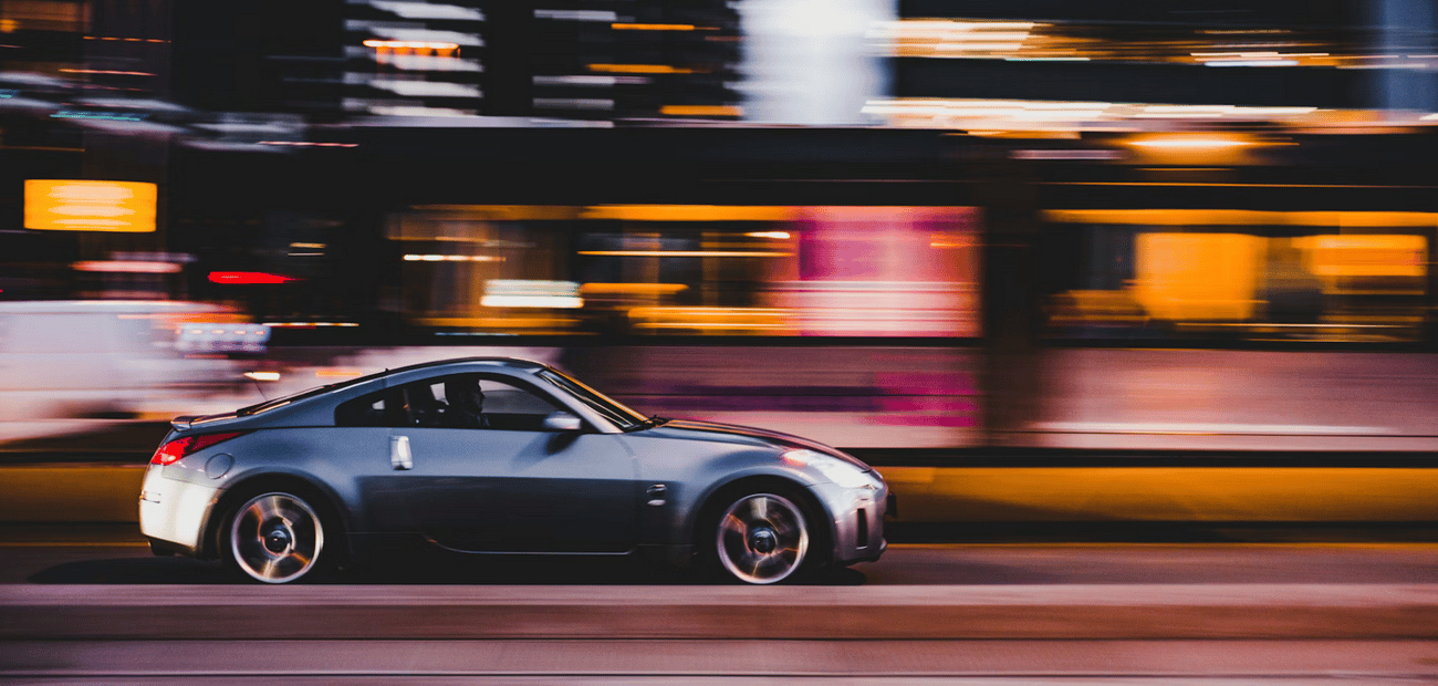 A silver sports car speeding through a city at night, the buildings and lights behind it blurred into streaks of orange and pink.