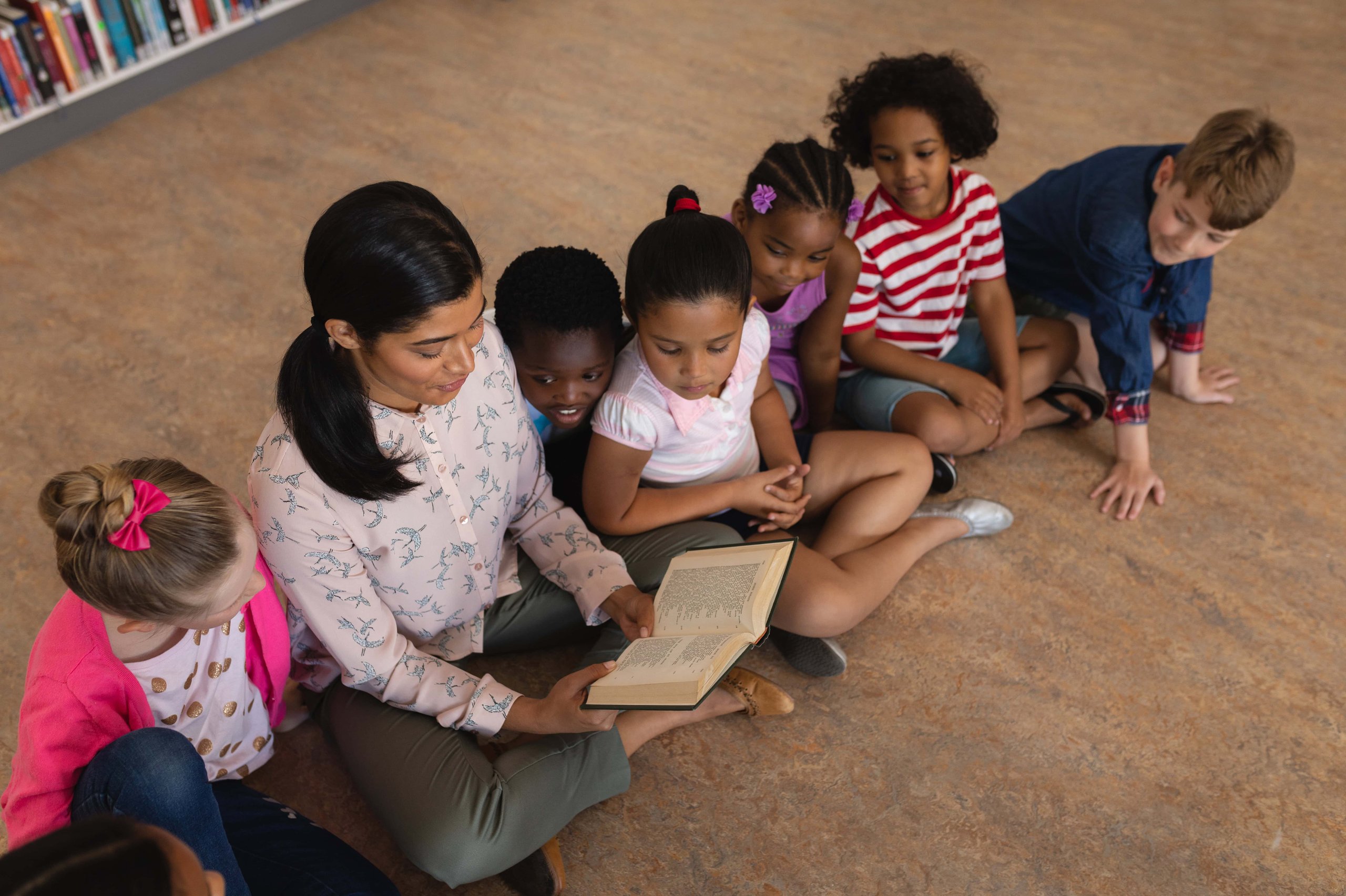 high angle view of female teacher teaching schoolkids and sitting on floor of school library