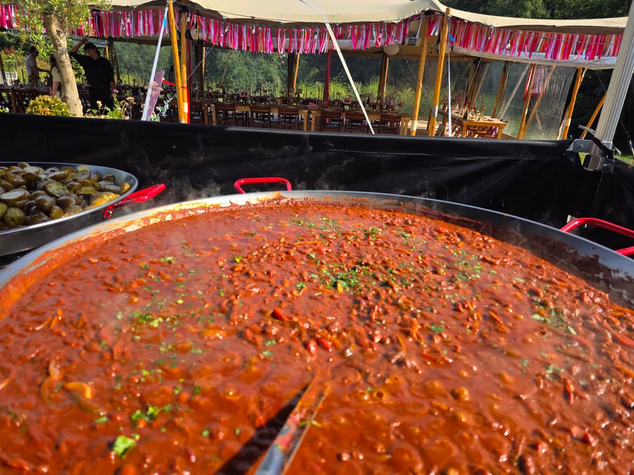 A large pan of simmering food, possibly a stew or curry, with another pan of roasted potatoes, under a marquee at an outdoor event