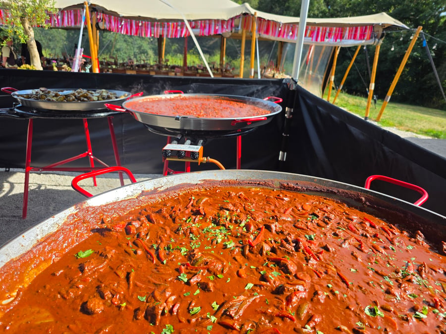 Three large paella pans are cooking various dishes, including a rich red stew and mussels, under a marquee at an outdoor event