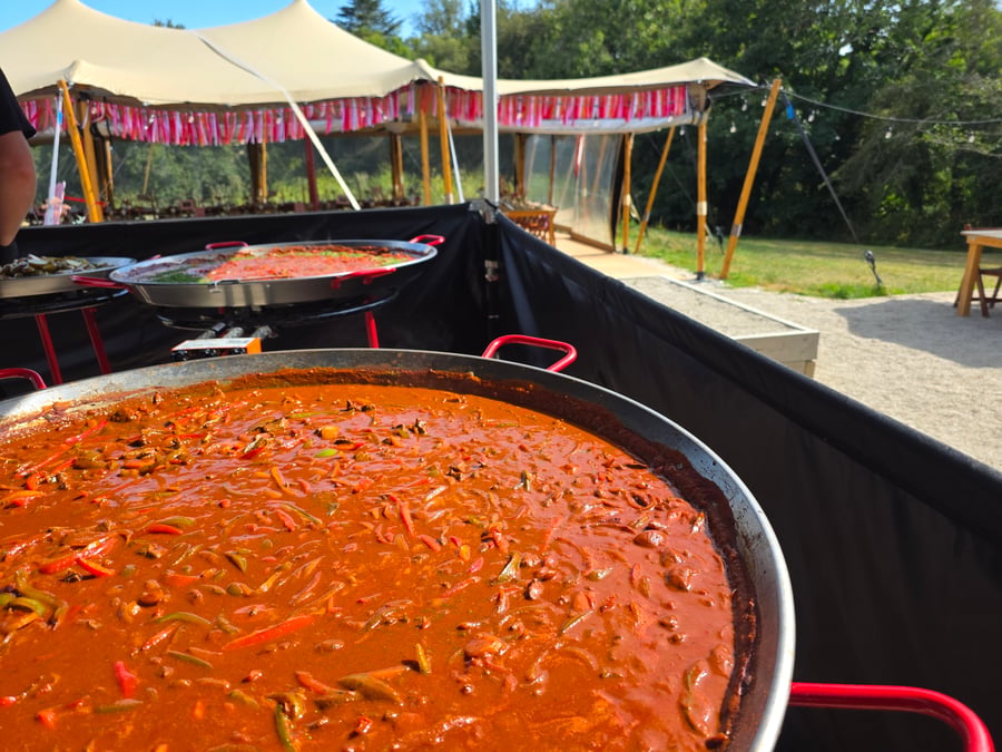 Two large paella pans filled with a rich, red dish are displayed at an outdoor catering event under a marquee