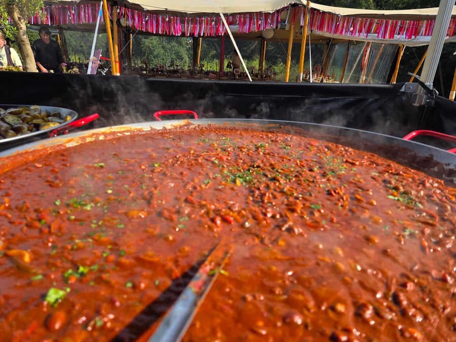 A large pan of steaming, rich red stew with green herbs is cooking outdoors, with a caterer visible in the background under a tent