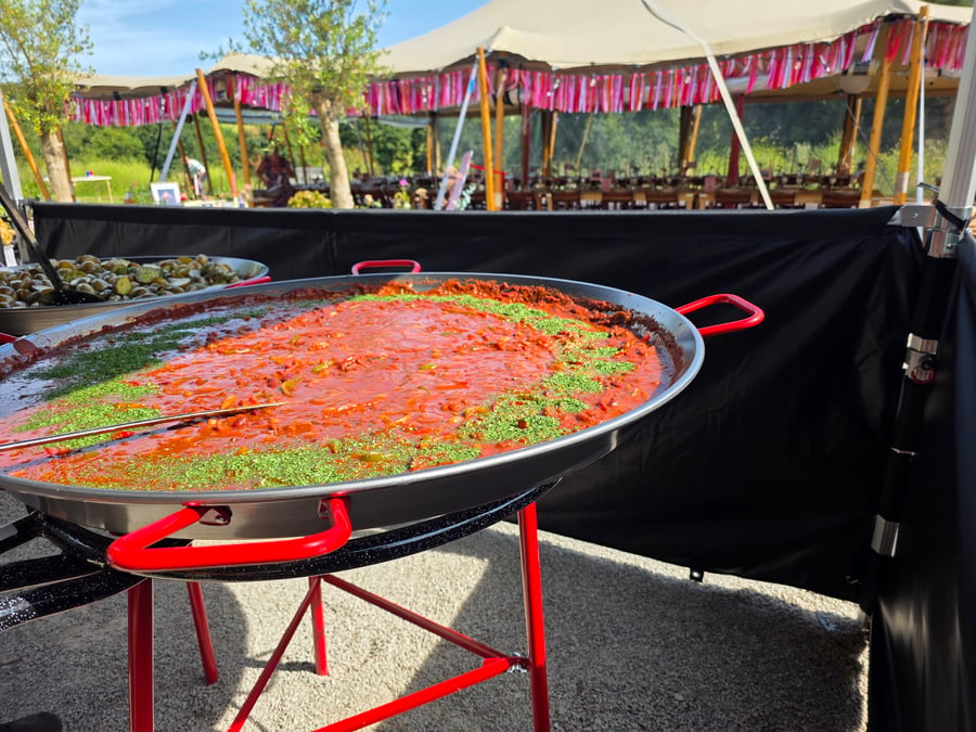 A large paella pan filled with a red sauce and green herbs is set up outdoors, with a marquee and tables visible in the background