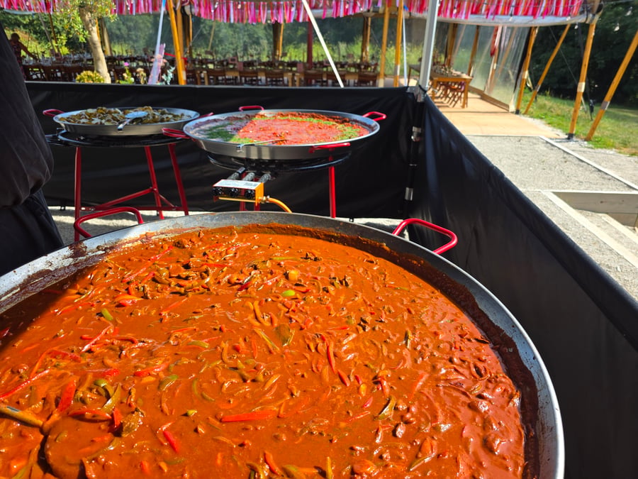 Three large pans of food, including a rich red stew and a green dish, are prepared outdoors for an event