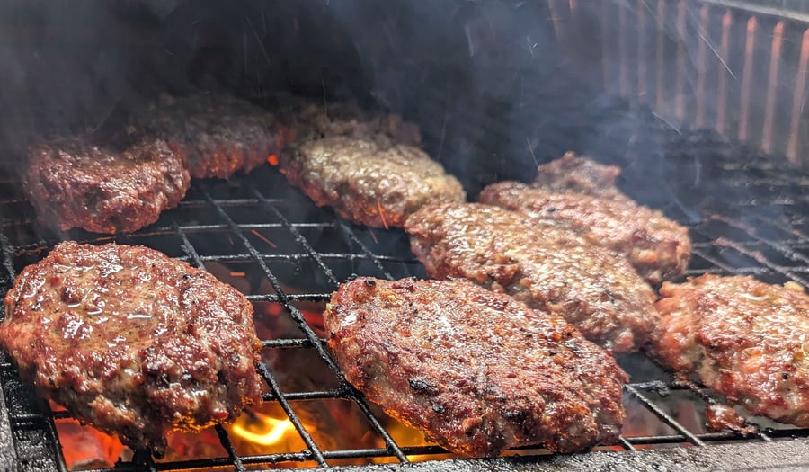 Juicy burgers sizzle on the grill over hot coals, ready for a delicious live-fire BBQ event