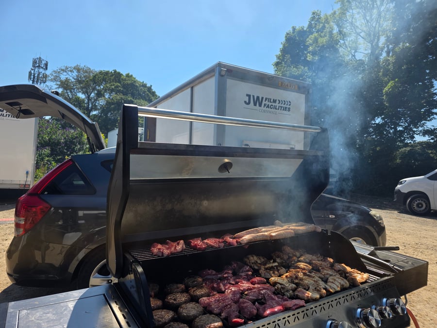 A large BBQ grill laden with burgers, sausages, and chicken cooking on a film set unit base in Cornwall