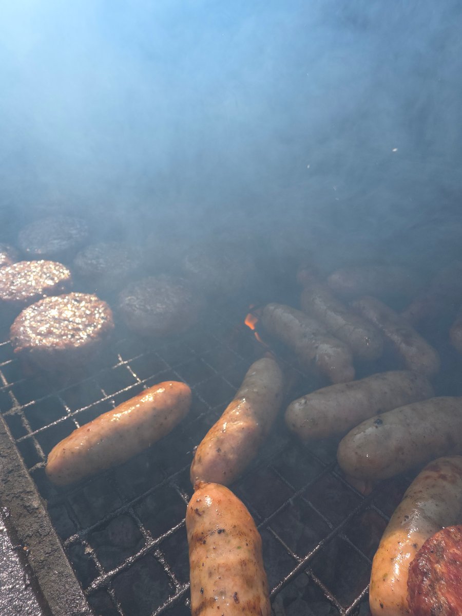 Sausages and burgers cooking on a grill, surrounded by smoke and a hint of flame, perfect for a live-fire event