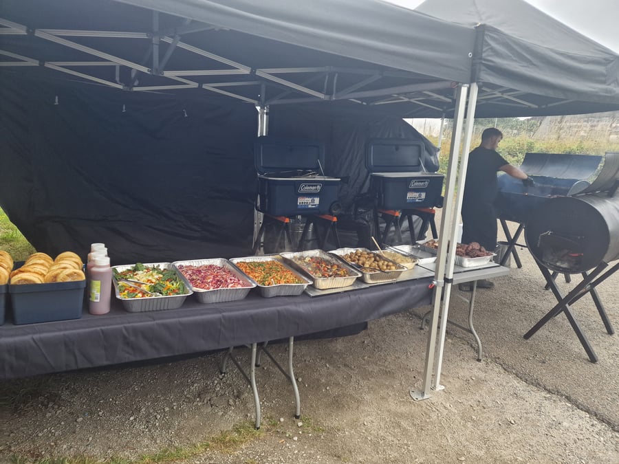 A chef prepares food on a large BBQ smoker next to a buffet spread of salads, potatoes, and bread rolls under a gazebo